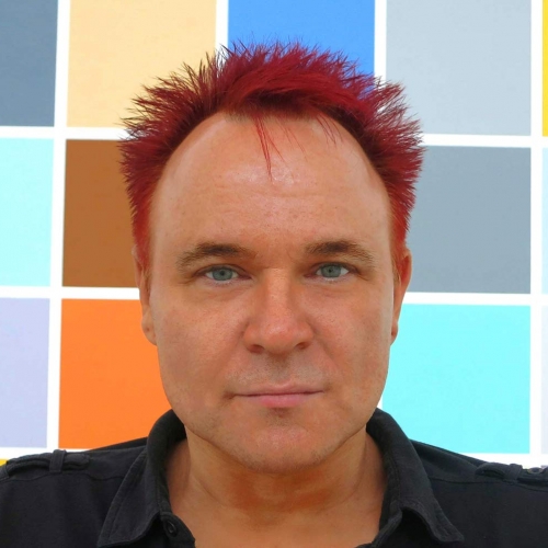 Mik looking directly into the camera, with red spikey hair and a coloured grid pattern behind him