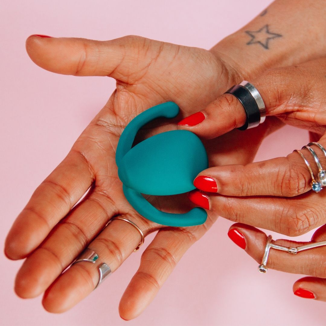 A woman holding a green vibrating sex toy. Her hands have tattoos, silver rings and red nail varnish
