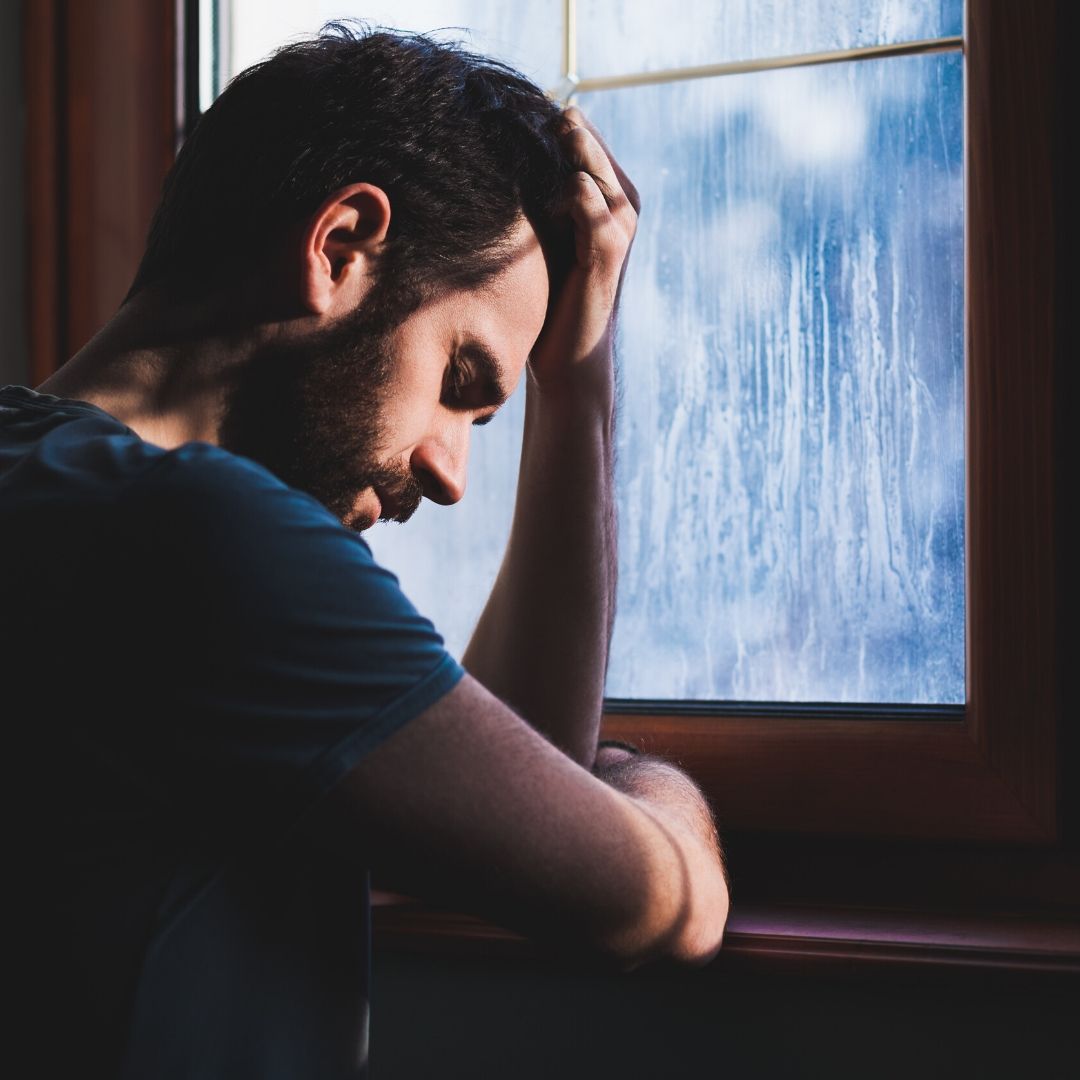A man with dark hair and a beard looks down with a sad expression whilst leaning against a window with rain on the pane