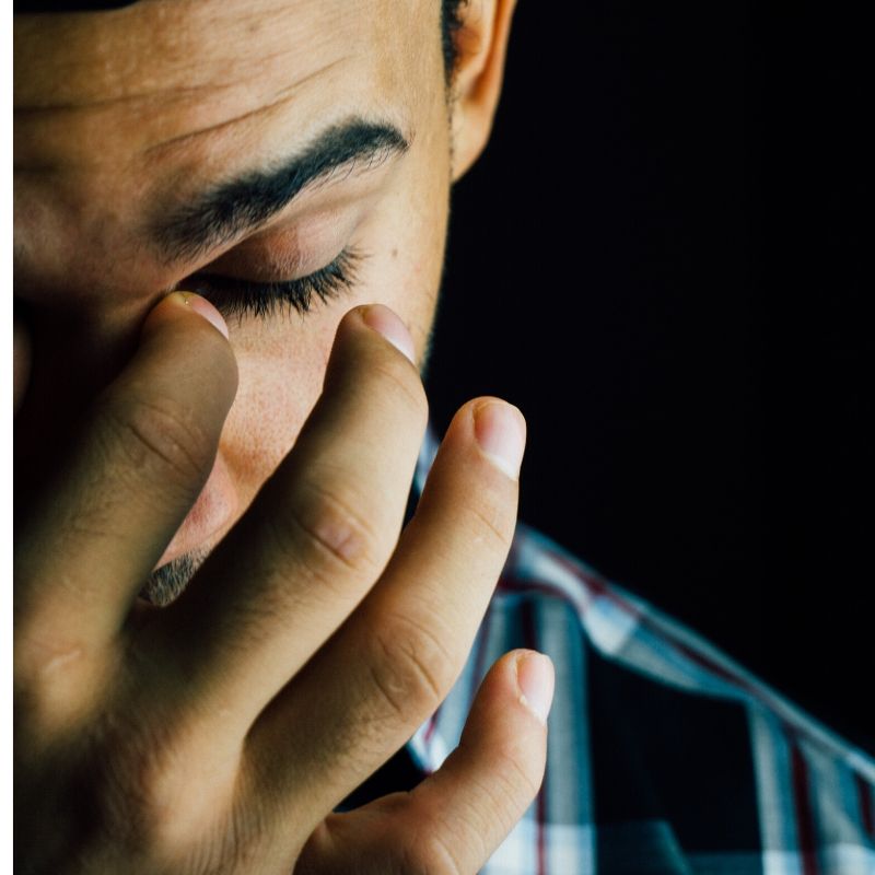 A man holds his hand to his face, his eye is closed and he looks down with a sad expression