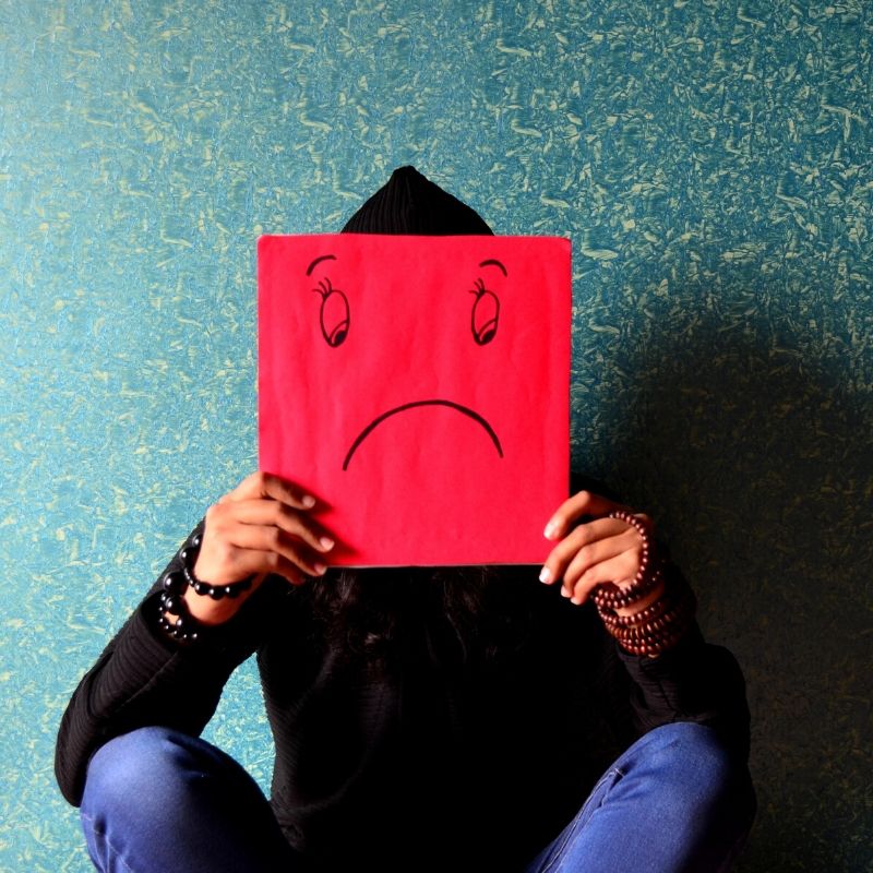 A man sitting down holding up a red square with a sad face drawn on it. He has brown skin, a black shirt, blue jeans and is wearing beaded bracelets. The background wall is turquoise blue