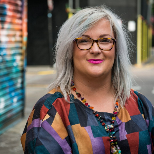 Emily Yates is photographed in the street. She is in her wheelchair and has grey blonde hair, tortoise shell glasses and vibrant pink lipstick. She's wearing a multicoloured top and a long beaded necklace.