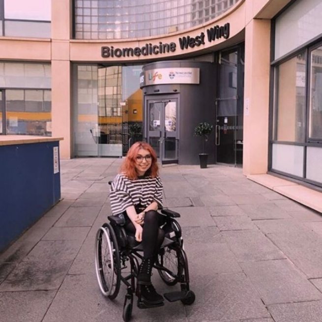 Georgia is sitting in her wheelchair in front of the hospital entrance. She's got dyed red hair, glasses and a big smile. She is wearing a baggy black and white striped t-shirt with black skirt and boots. Biomedicine West Wing is written in letters above the hospital entrance.