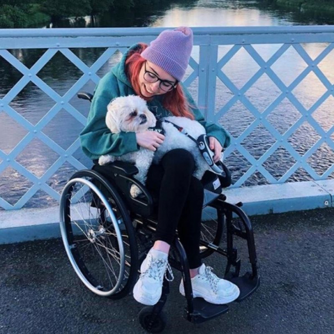 Georgia is cuddling her white dog on her knee. She is on a bridge with blue criss cross railings and the river behind her. She's wrapped up for the cold in a lilac beanie hat and green hoodie.