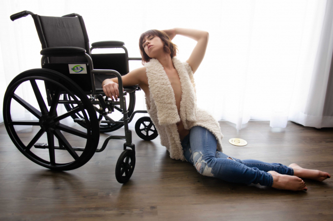 Josie is wearing jeans and a white sleeveless open top, revealing her bare arms and part of her chest. She is sitting on a wooden floor with her back and arm resting against her wheelchair.