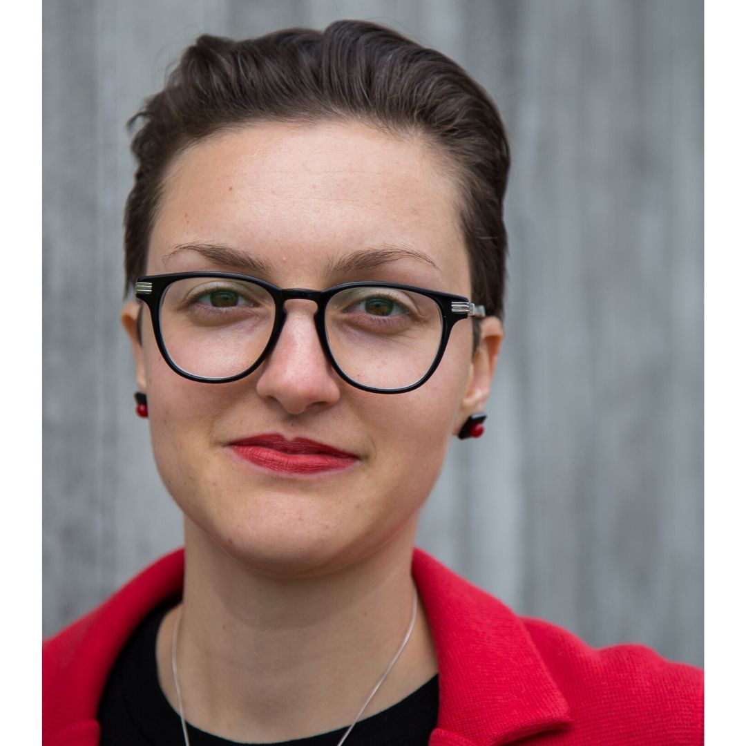 Julia Bahner wears a red suit jacket and a black top. She has short dark hair, red lipstick and glasses.
