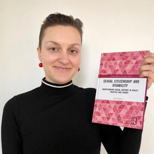 Julia is pictured wearing a black polo neck jumper and red drop earrings. She is holding a copy of her book, which has pink hexagons on the cover. She has short dark brown hair and is smiling at the camera.